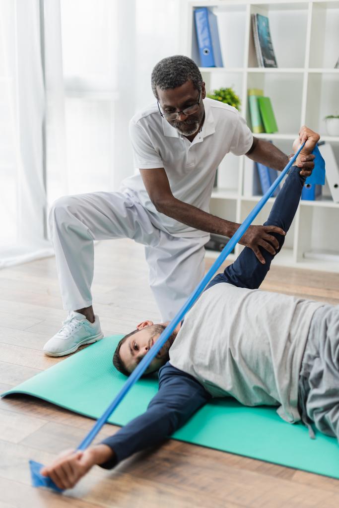 rehabilitólogo afroamericano maduro ayudando a un joven a entrenar con una banda elástica en una esterilla de fitness - Foto, Imagen