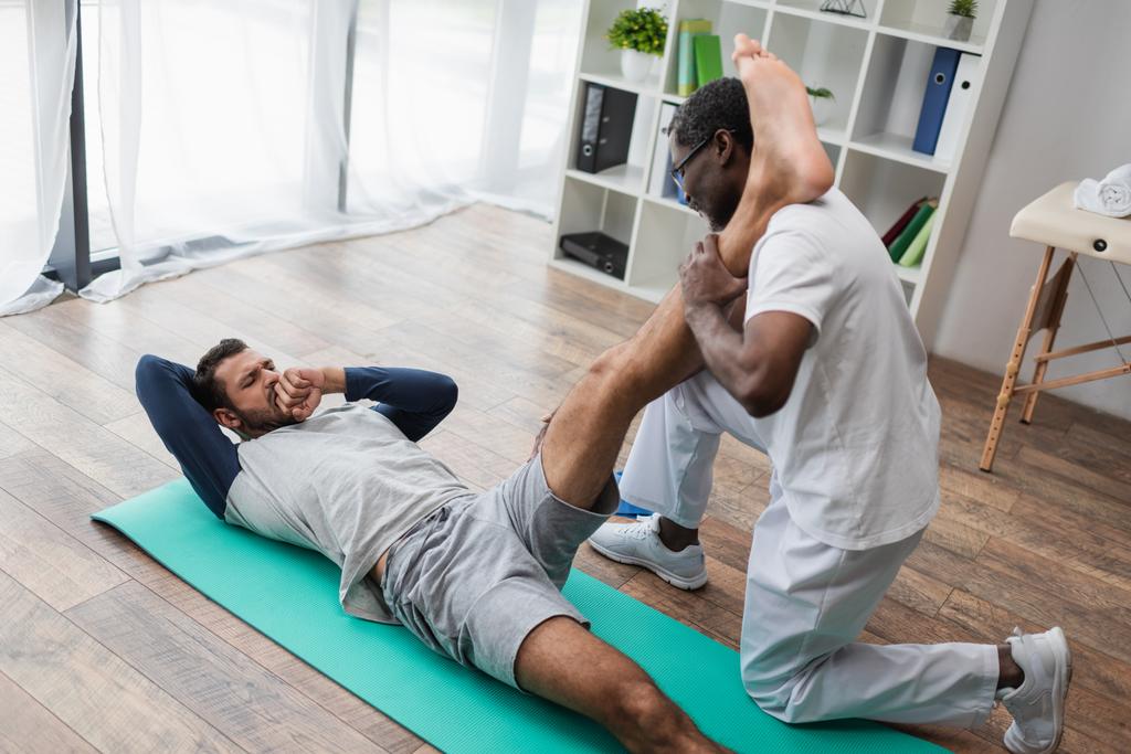 Afrikalı Amerikalı rehabilitasyon uzmanı bacağını esnetirken genç adam acı çekiyor. - Fotoğraf, Görsel