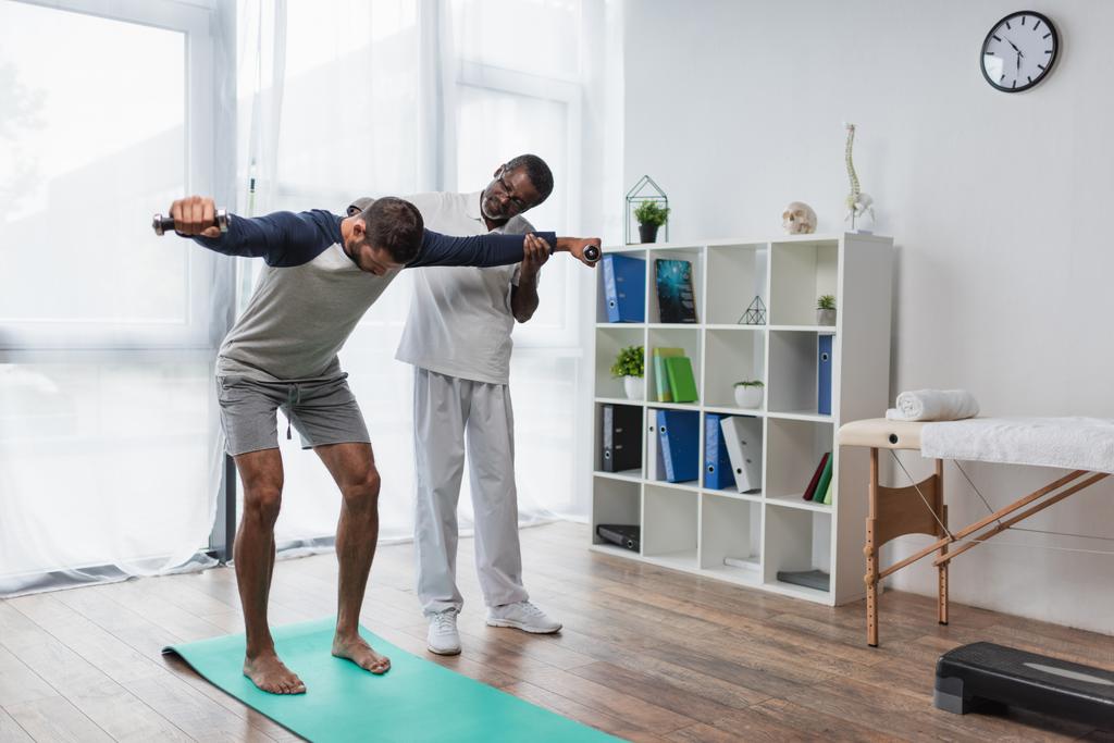 érett afro-amerikai rehabilitológus segít a fiatal férfi edzés súlyzók fitness mat - Fotó, kép