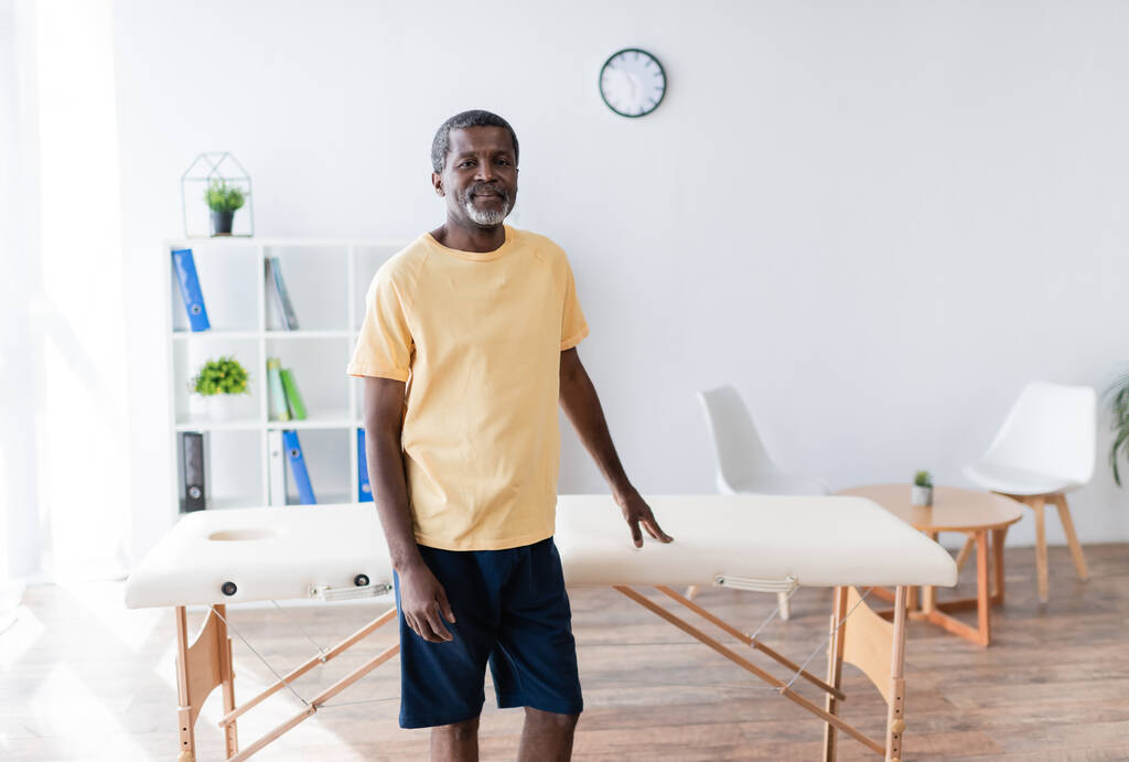 позитивний афроамериканський чоловік стоїть біля масажного столу в реабілітаційному центрі і дивиться на камеру
 - Фото, зображення
