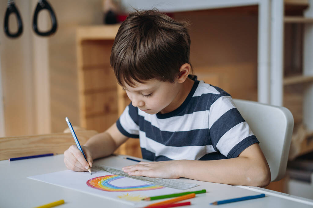 adorable chico caucásico de edad elemental dibujando un arco iris con lápices sentados en el escritorio de su habitación en casa. Foto de alta calidad - Foto, Imagen