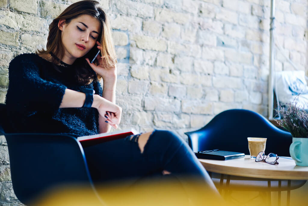 Młody zdalny pracownik w ubraniu siedzącym na krześle w kawiarni z notebookiem i kawą podczas rozmowy telefonicznej i sprawdzania informacji o zegarku - Zdjęcie, obraz