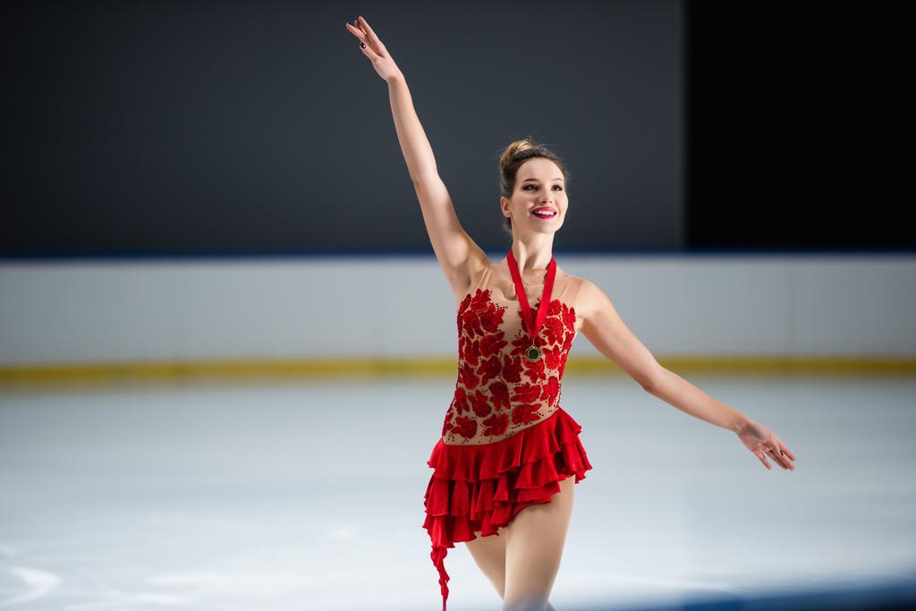 positieve kunstschaatser in rode jurk en gouden medaille gebaar op de ijsbaan - Foto, afbeelding