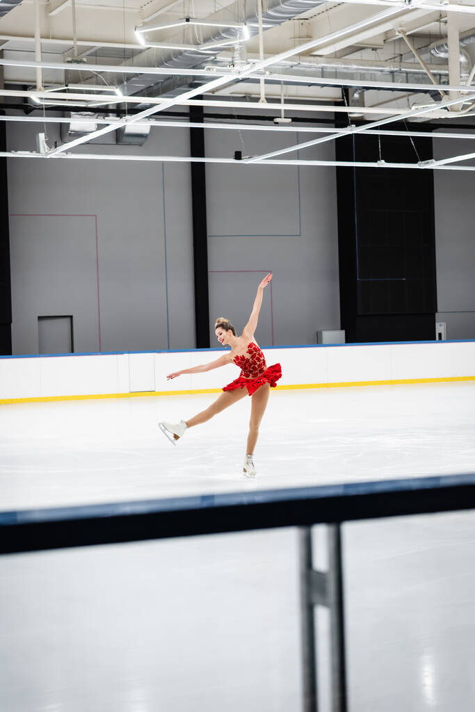 pleine longueur de femme heureuse en robe rouge patinage artistique avec les mains tendues dans l'arène de glace professionnelle - Photo, image