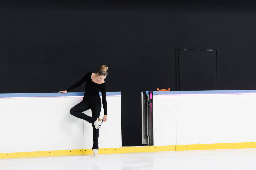 volledige lengte van de kunstschaatser in zwart bodysuit controleren mes op schaatsen in de buurt van bevroren ijs arena - Foto, afbeelding
