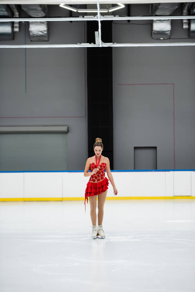 longitud completa de joven patinador artístico en vestido rojo mirando medalla de oro en pista de hielo - Foto, imagen