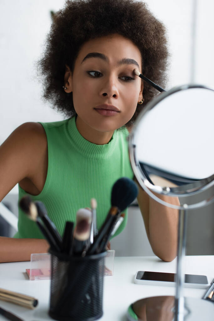 Mujer afroamericana aplicando sombra de ojos cerca de espejo y smartphone  - Foto, imagen