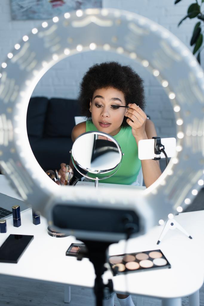 Blogueur afro-américain appliquant mascara près de smartphone et anneau lumineux à la maison  - Photo, image