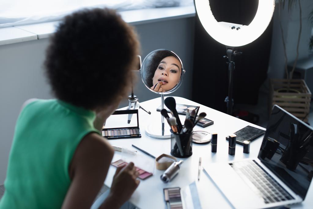 Blurred african american blogger applying decorative cosmetics near gadgets and ring light at home  - Foto, Bild