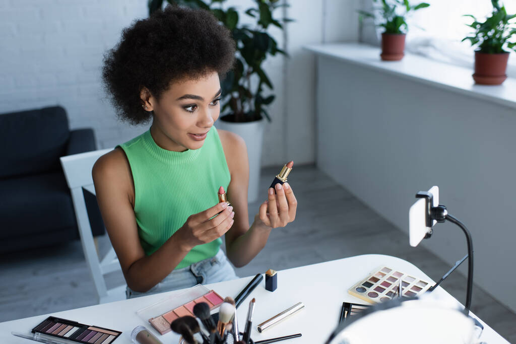 Afrykański amerykański bloger trzyma szminki w pobliżu smartfona w domu  - Zdjęcie, obraz