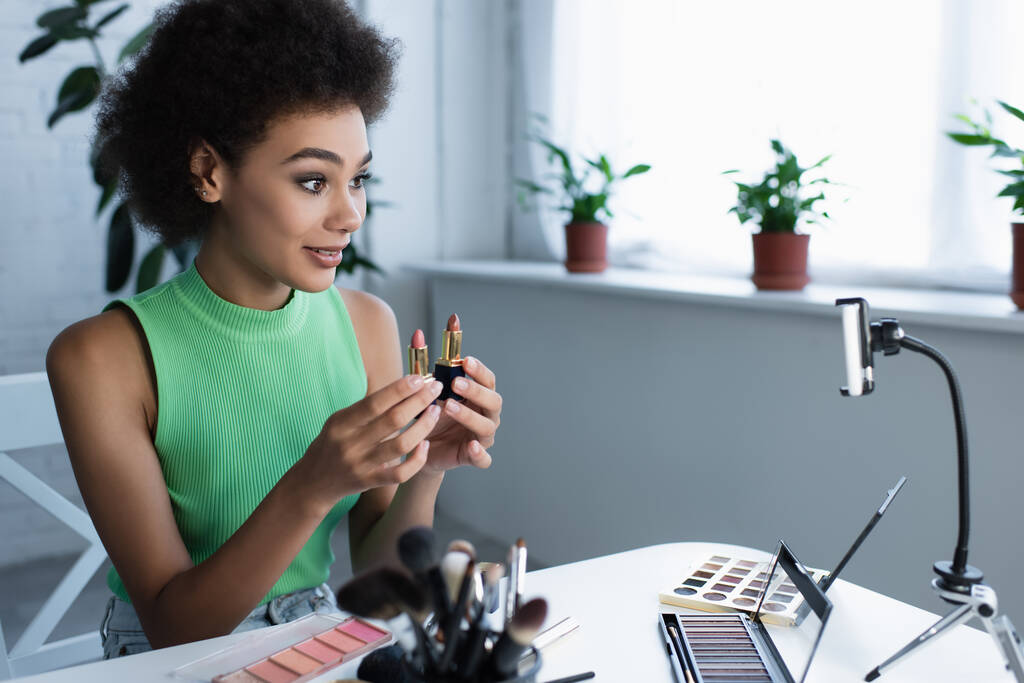 blogger afroamericano sorridente che tiene rossetti vicino a cosmetici decorativi e smartphone  - Foto, immagini