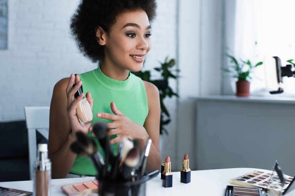 Femme afro-américaine positive montrant fond de teint près des cosmétiques à la maison  - Photo, image