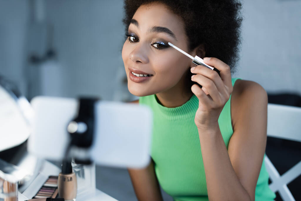Positive african american blogger looking at smartphone while applying eye shadows  - Foto, Imagem