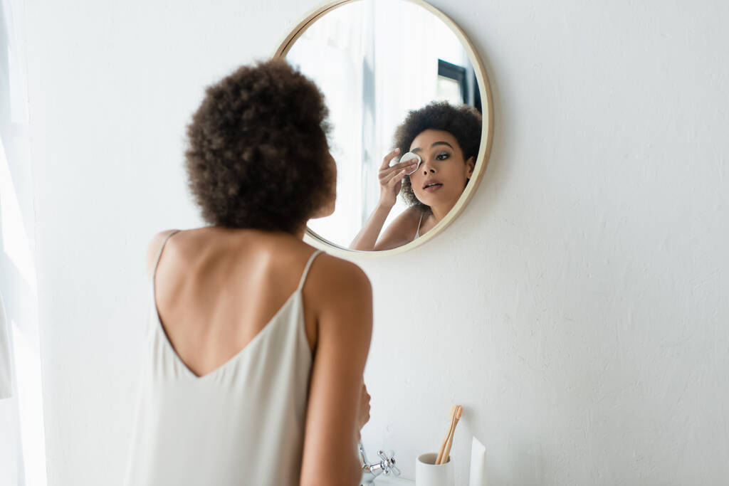 Joven mujer afroamericana en camisón de seda limpiando el ojo con almohadilla de algodón cerca del espejo en el baño  - Foto, imagen