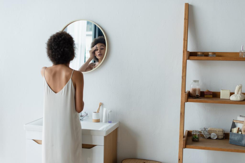 Молода афроамериканська жінка в атласній нічній сукні витирає обличчя бавовняною подушкою біля дзеркала і туалетів у ванній кімнаті.  - Фото, зображення
