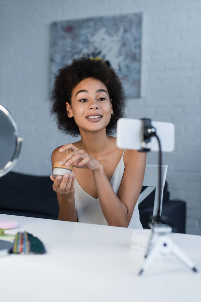 Sonriente blogger afroamericano sosteniendo crema cosmética cerca de teléfono inteligente y espejo en casa  - Foto, imagen