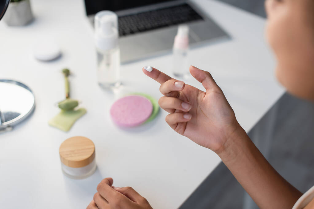 Cropped view of african american woman holding cosmetic cream near blurred laptop and jade roller on table  - Photo, Image