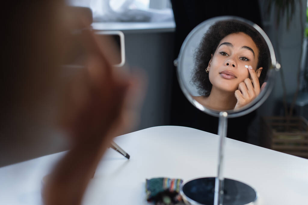 Afrikaanse amerikaanse blogger aanbrengen van cosmetische crème op gezicht in de buurt van spiegel en smartphone thuis  - Foto, afbeelding