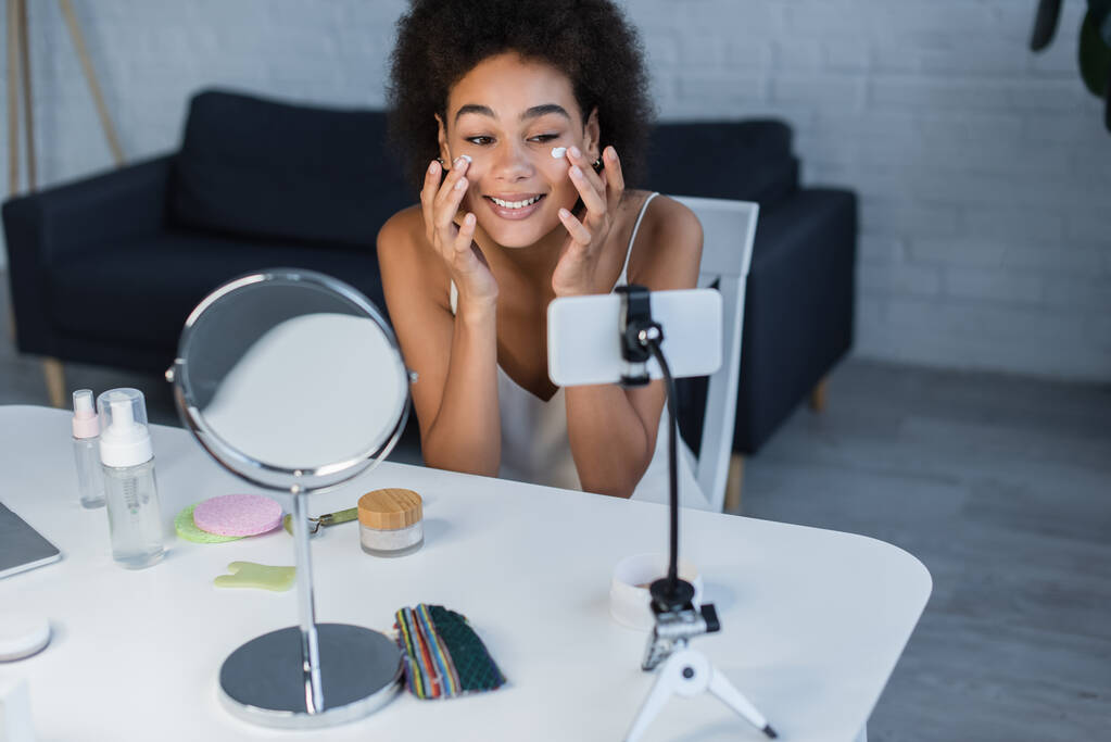 Allegro blogger afroamericano che applica crema cosmetica vicino al cellulare e rullo di giada a casa  - Foto, immagini