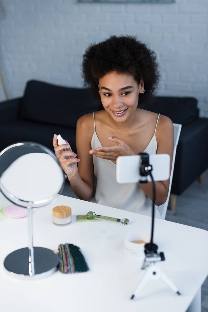 Улыбающийся африканский американский блогер указывает на косметический лосьон рядом со смартфоном и зеркалом дома  - Фото, изображение
