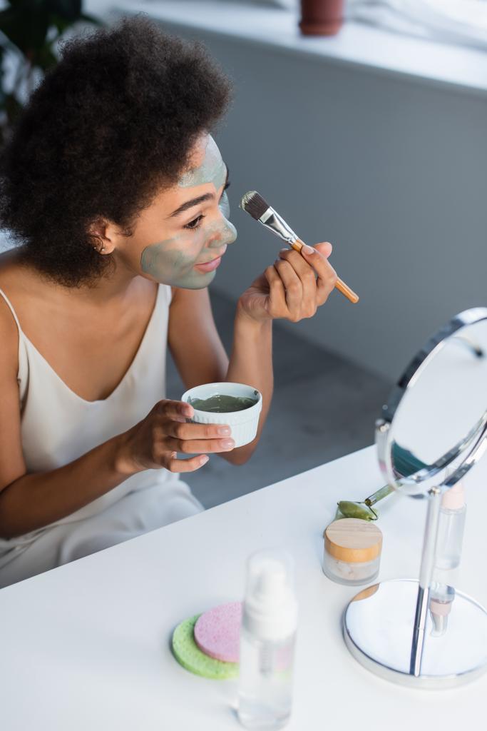 Mujer afroamericana aplicando mascarilla de arcilla cerca de crema cosmética, rodillo de jade y espejo en casa  - Foto, Imagen