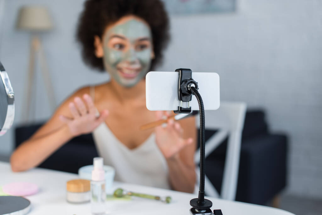 Smartphone v blízkosti rozmazané africký americký blogger s hliněnou maskou na tváři doma  - Fotografie, Obrázek