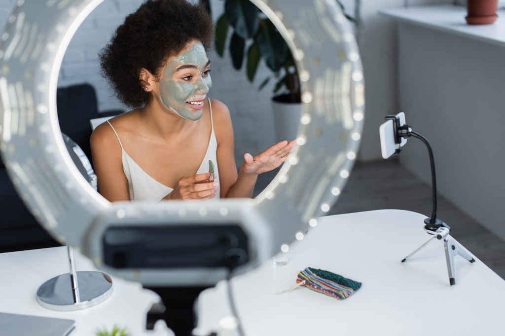 Blogger afroamericano sonriente con máscara de arcilla en la cara sosteniendo cepillo cosmético cerca de teléfono inteligente y anillo de luz  - Foto, Imagen