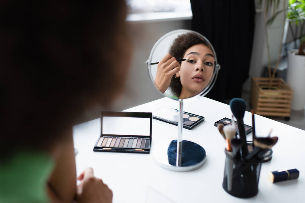 Wazig Afro-Amerikaanse vrouw aanbrengen oogschaduw in de buurt van spiegel en cosmetische borstels thuis  - Foto, afbeelding