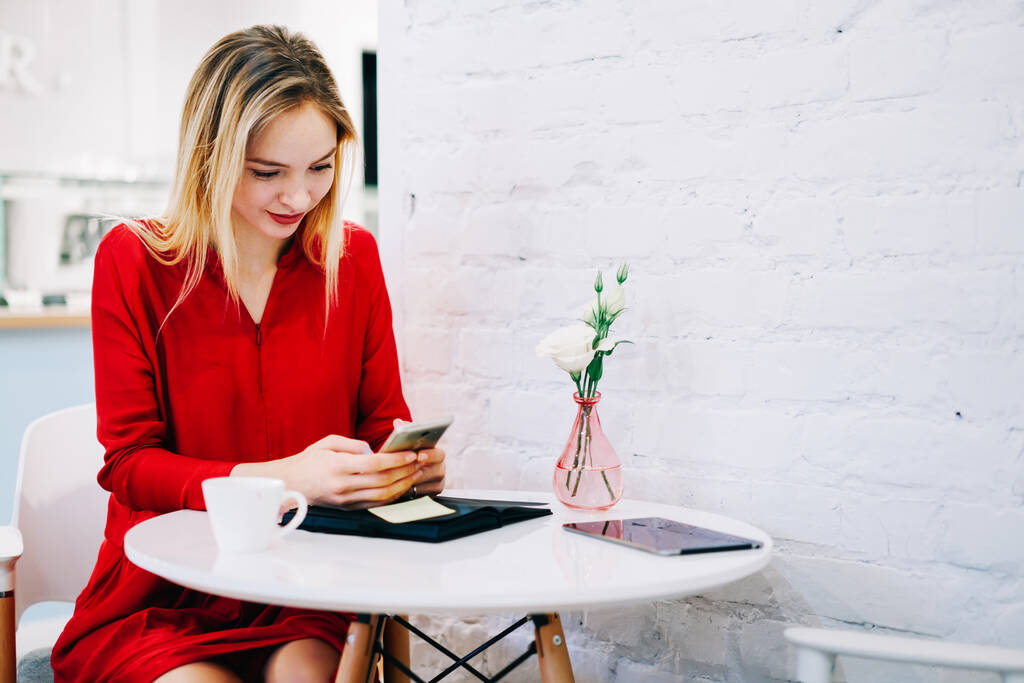 Fiatal hölgy szőke haj öltözött piros ruha ül asztalnál hangulatos kávézó, miközben szörfözés okostelefon és ivás csésze kávét - Fotó, kép