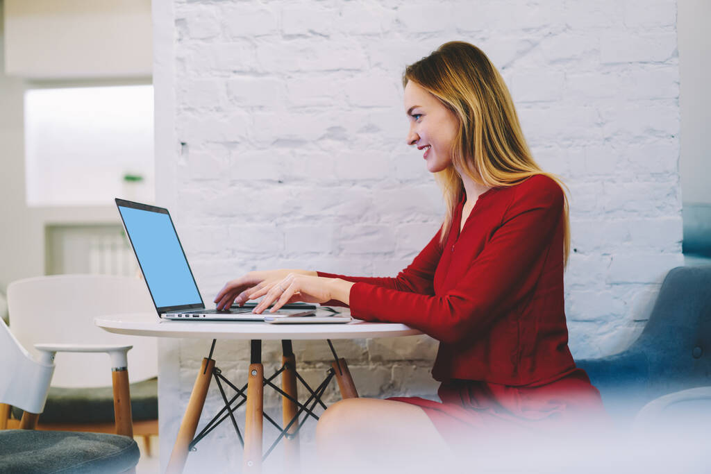 Вид збоку весела жінка в стильному одязі, сидячи за столом і друкуючи на ноутбуці під час роботи над проектом в кафе
 - Фото, зображення