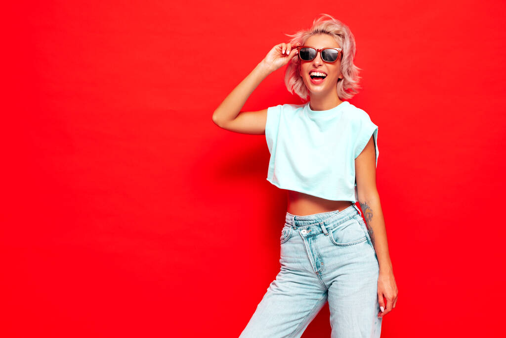 Jeune belle femme souriante en vêtements d'été à la mode. Femme sexy insouciante posant près du mur rouge en studio. Modèle blond positif s'amuser et devenir fou. Joyeux et heureux. Dans les lunettes de soleil - Photo, image