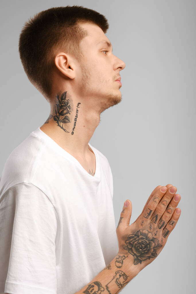 Νεαρός άνδρας με τατουάζ στέκεται στο προφίλ, κρατά τις παλάμες των χεριών του ενωμένες και προσεύχεται - Φωτογραφία, εικόνα