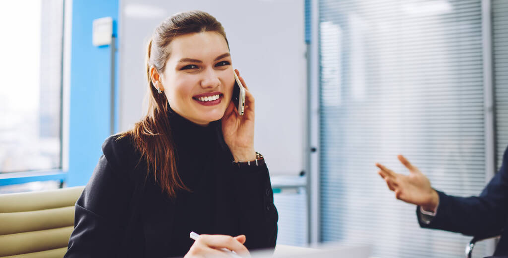 Cheerful adult businesswoman wearing black turtleneck sitting with colleagues in contemporary light office and talking on mobile phone while smiling at camera - Photo, Image