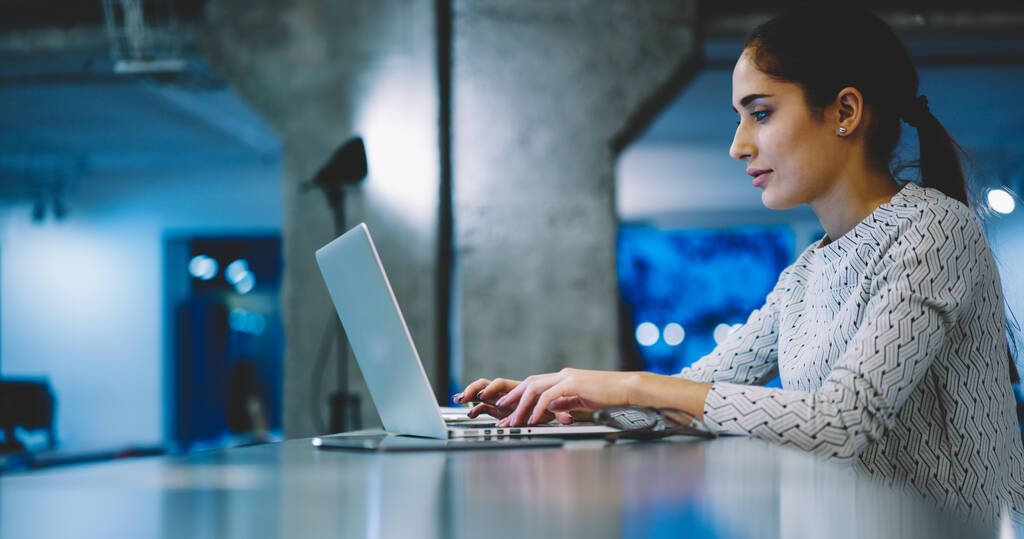 Seitenansicht einer konzentrierten Frau in lässigem Outfit, die am Netbook tippt, während sie am Schreibtisch in einem modernen Arbeitsbereich an einem neuen Projekt arbeitet - Foto, Bild