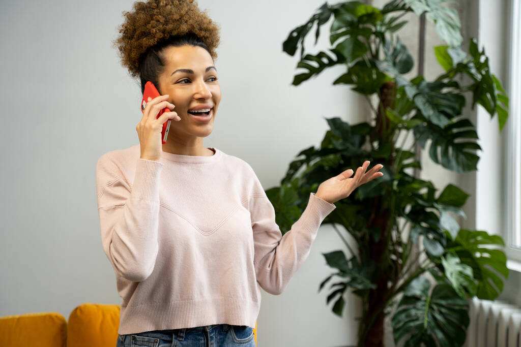Egy fiatal afro-amerikai nő beszél telefonon, a nappaliban áll. Egy üzletasszony, aki egy otthoni irodában dolgozik és mobiltelefont használ.. - Fotó, kép