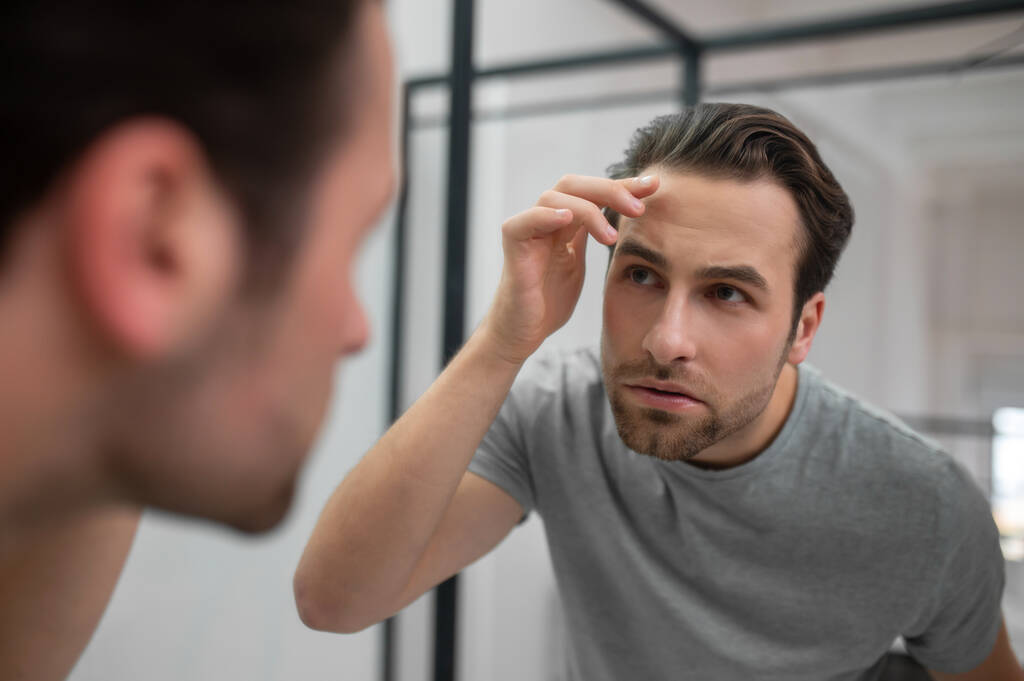 Een jongeman in grijs T-shirt kijkt naar zijn spiegelbeeld in de spiegel - Foto, afbeelding