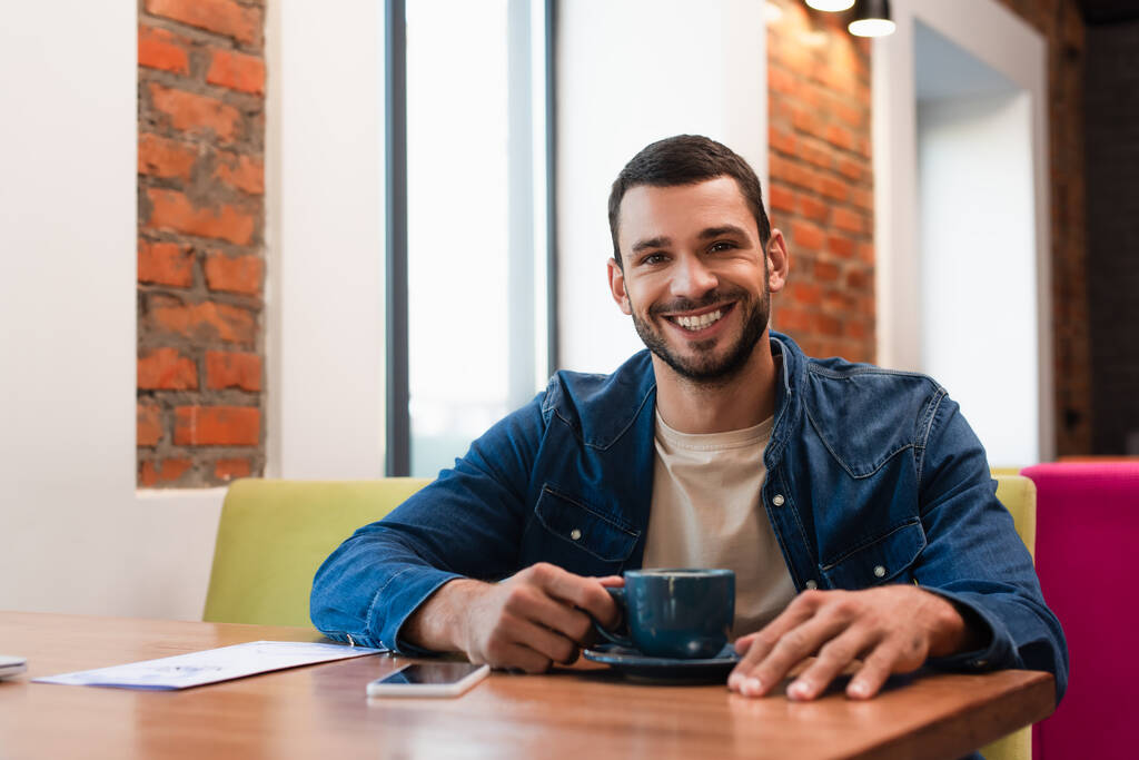 homem alegre olhando para a câmera perto da xícara de café e smartphone com tela em branco no café - Foto, Imagem