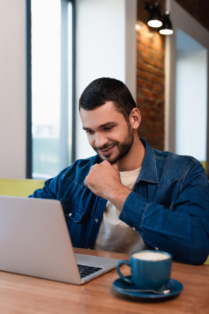 χαμογελαστός άνθρωπος κοιτάζοντας φορητό υπολογιστή κοντά θολή φλιτζάνι καφέ στο καφέ - Φωτογραφία, εικόνα