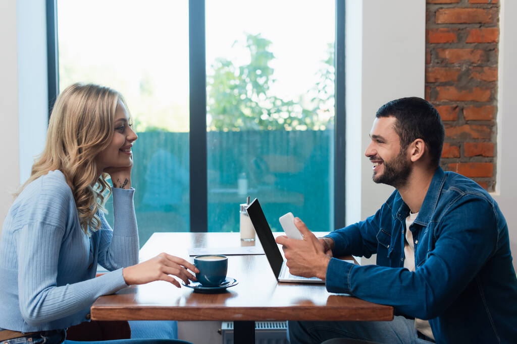Seitenansicht eines glücklichen Paares, das sich in der Nähe eines Laptops mit leerem Bildschirm und einer Tasse Cappuccino anschaut  - Foto, Bild