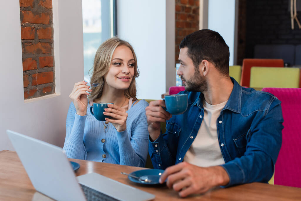 happy couple with coffee cups looking at each other near blurred laptop in restaurant - Photo, Image