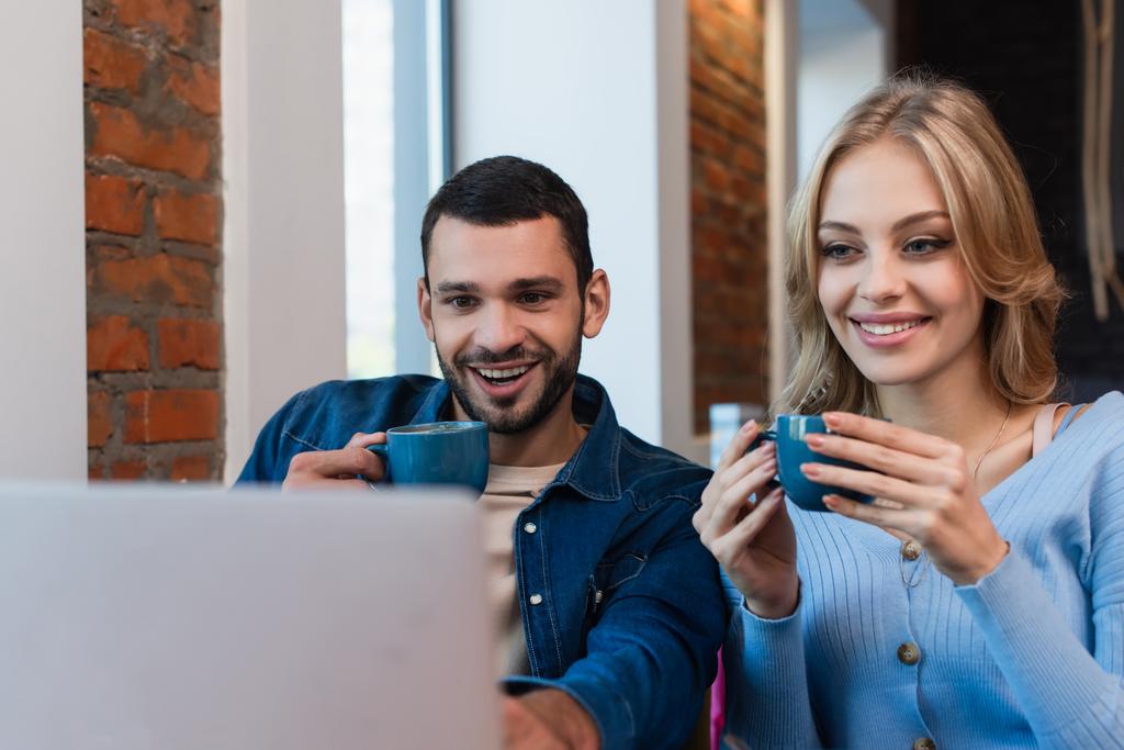 fröhliches Paar mit Kaffeetassen, das auf verschwommenen Laptop schaut, während es Zeit im Café verbringt - Foto, Bild