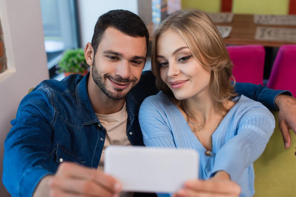 pareja complacida tomando selfie en el teléfono celular borroso mientras pasa tiempo en la cafetería - Foto, imagen