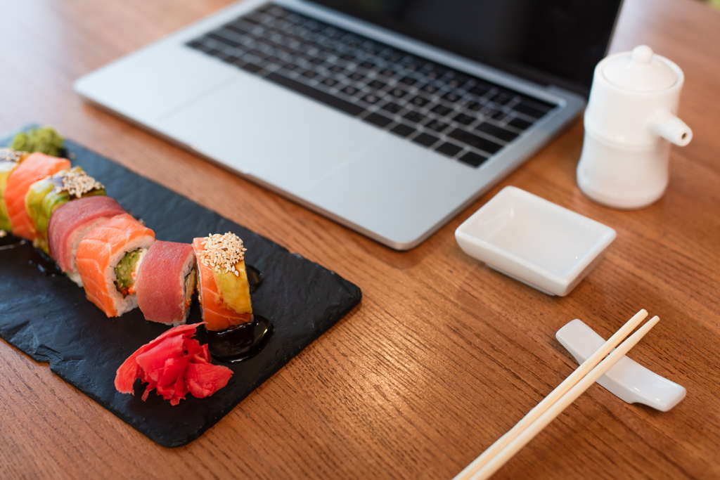 savoureux rouleaux de sushi, baguettes, pot de sauce soja et bol près ordinateur portable flou sur la table dans le bar à sushi - Photo, image