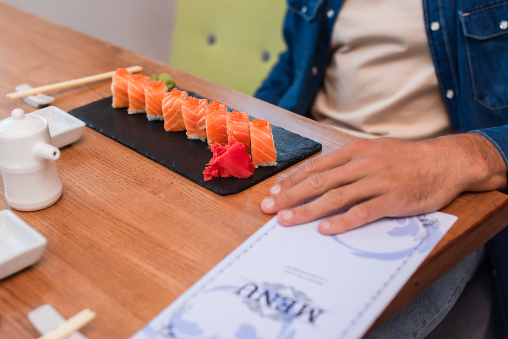 bijgesneden beeld van de mens in de buurt plaat van sushi rollen en wazig menu op tafel - Foto, afbeelding