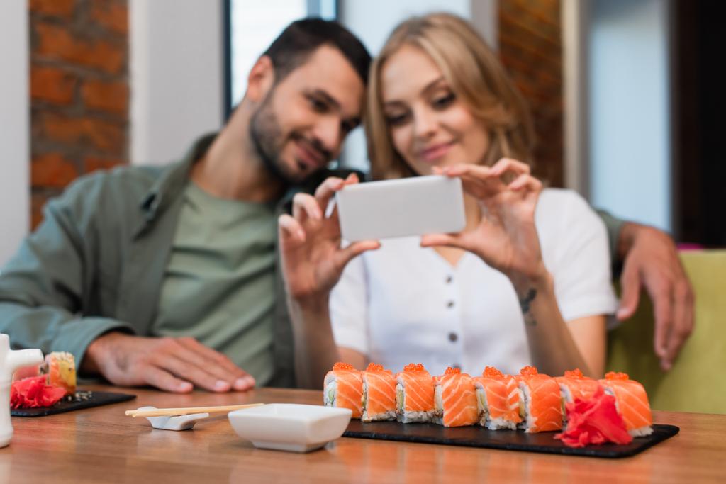 blurred woman taking photo of sushi rolls near happy boyfriend in sushi bar - Photo, Image