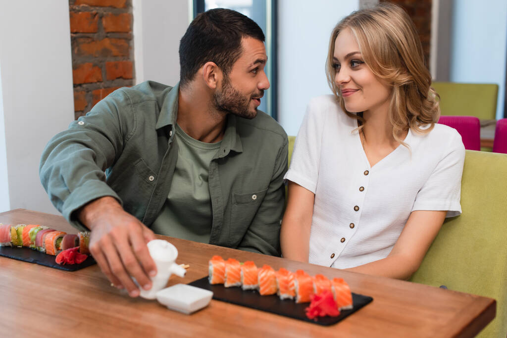 aufgeregter Mann spricht mit lächelnder Freundin, während er einen Sojasauce-Topf in der Nähe von Sushi-Brötchen hält - Foto, Bild