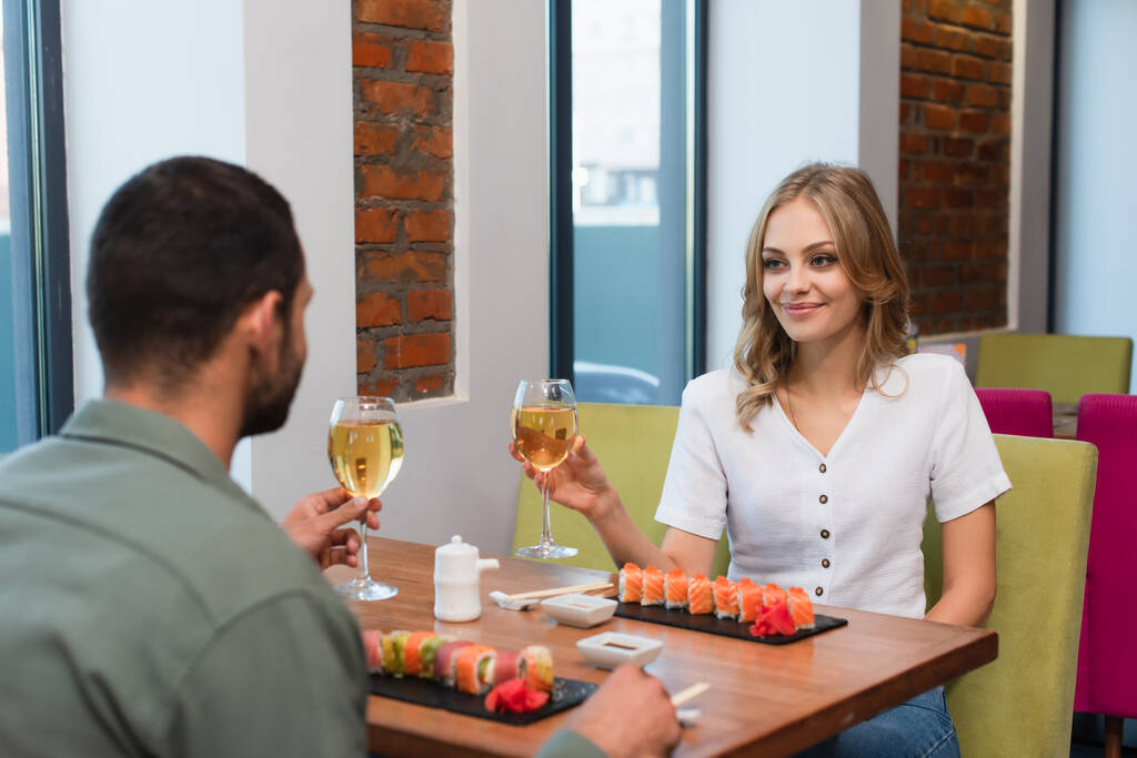 νεαρό ζευγάρι κρατώντας ποτήρια λευκό κρασί κοντά σε νόστιμα ρολά σούσι στο εστιατόριο - Φωτογραφία, εικόνα
