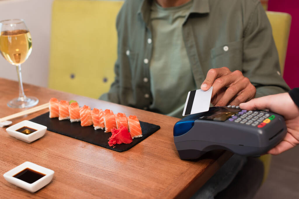 vista recortada del hombre borroso que paga a través de lector de tarjetas de crédito cerca de rollos de sushi y copa de vino - Foto, Imagen