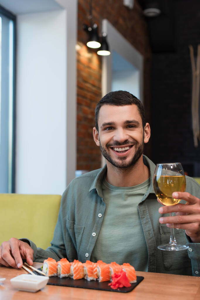 Bir bardak beyaz şarapla mutlu bir adam elinde lezzetli suşiyle tabağın yanındaki kameraya bakıyor. - Fotoğraf, Görsel