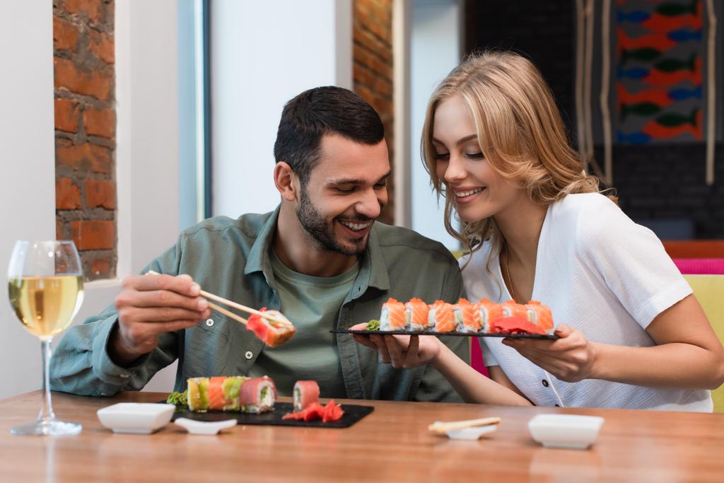 χαμογελαστή γυναίκα δείχνει νόστιμα ρολά σούσι στο φίλο κατά τη διάρκεια του δείπνου στο εστιατόριο - Φωτογραφία, εικόνα
